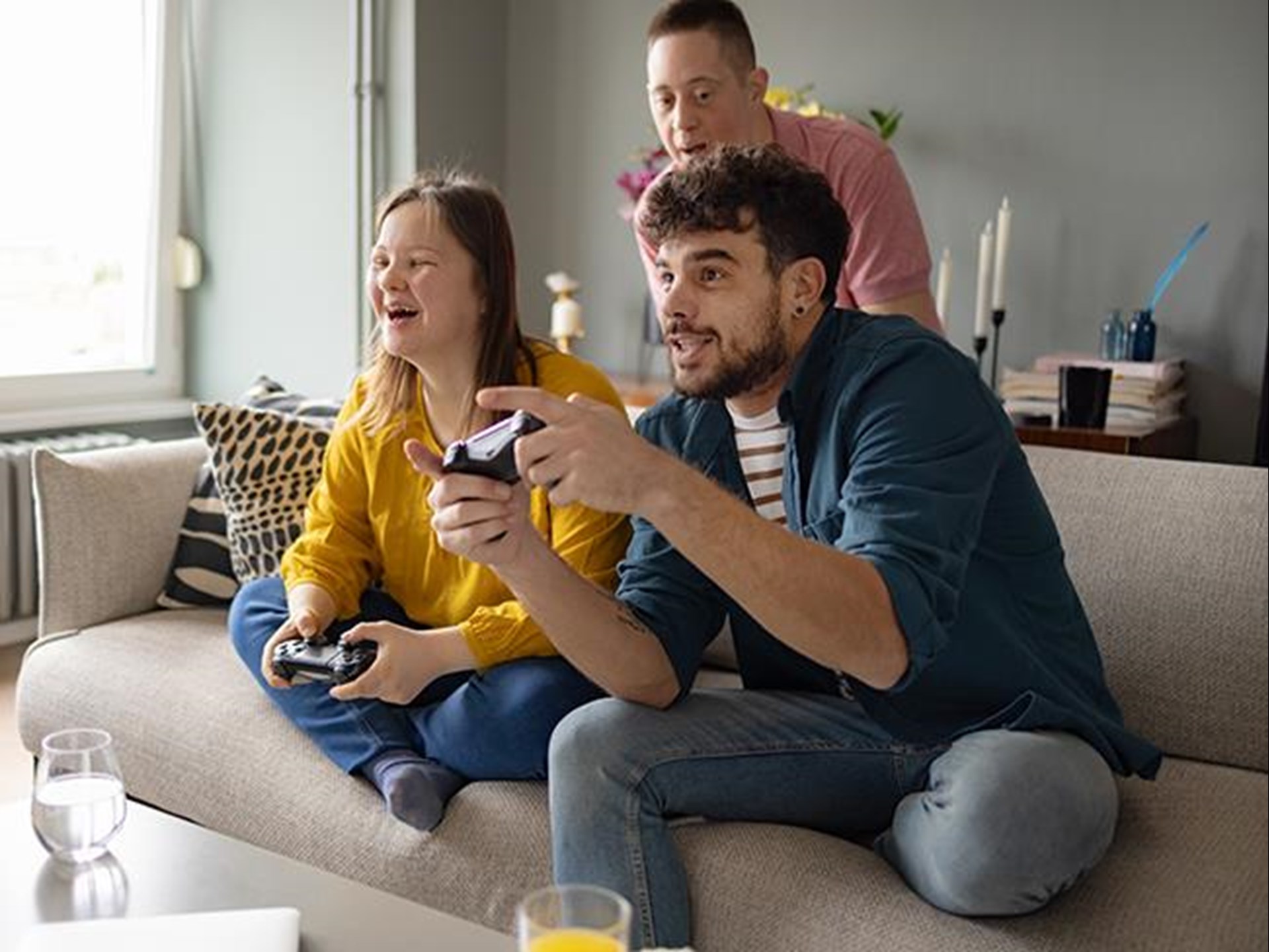 Eine junge Frau und ein junger Mann sitzen auf der Couch. Sie beide halten je einen Controller in der Hand. Der junge Mann trägt einen Bart. Er beteiligt sich an dem Computerspiel mit voller Leidenschaft. Dieser junge Mann ist auch der Assistent der jungen Frau. Sie hat das Downsyndrom. Ein weiterer junger Mann - ebenfalls mit Downsyndrom steht hinter der Couch. Er und die junge Frau, mit der er in der Wohnung lebt, lachen über den Spieleifer des Assistenten. 