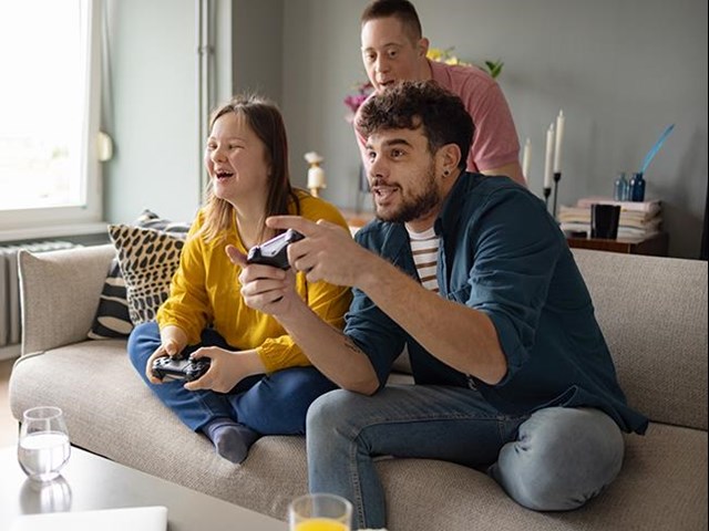 Eine junge Frau und ein junger Mann sitzen auf der Couch. Sie beide halten je einen Controller in der Hand. Der junge Mann trägt einen Bart. Er beteiligt sich an dem Computerspiel mit voller Leidenschaft. Dieser junge Mann ist auch der Assistent der jungen Frau. Sie hat das Downsyndrom. Ein weiterer junger Mann - ebenfalls mit Downsyndrom steht hinter der Couch. Er und die junge Frau, mit der er in der Wohnung lebt, lachen über den Spieleifer des Assistenten. 