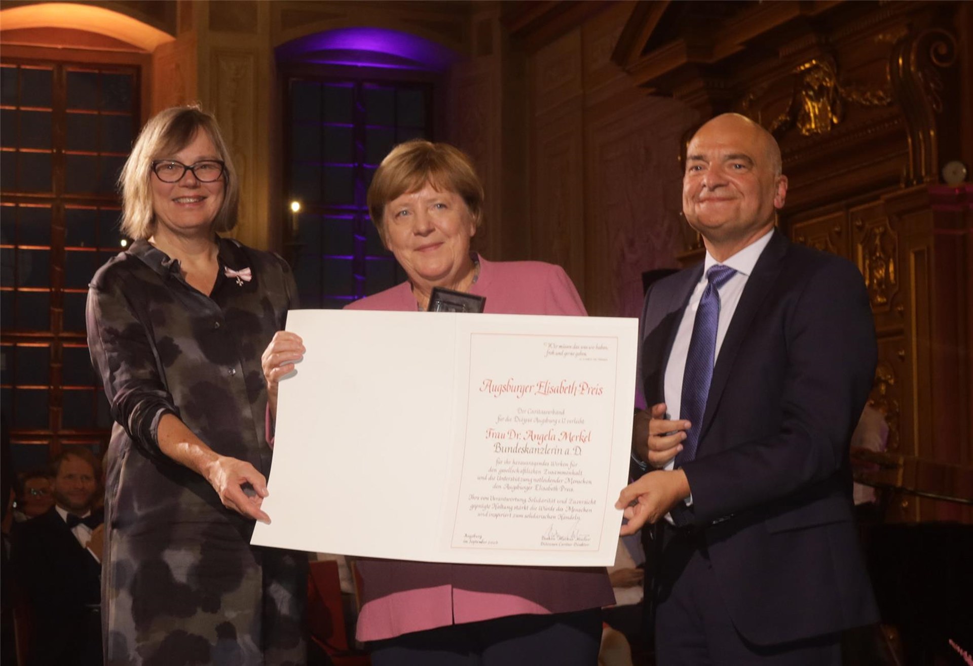 Das Foto zeigt Bundeskanzlerin a. D. Dr. Angela Merkel zwischen der Caritas-Präsidentin Eva Maria Welskop-Deffaa (links) und dem Augsburger Diözesan-Caritasdirektor Diakon Markus Müller. Merkel hält dabei die große Urkunde des Elisabeth-Preises in der Hand. 