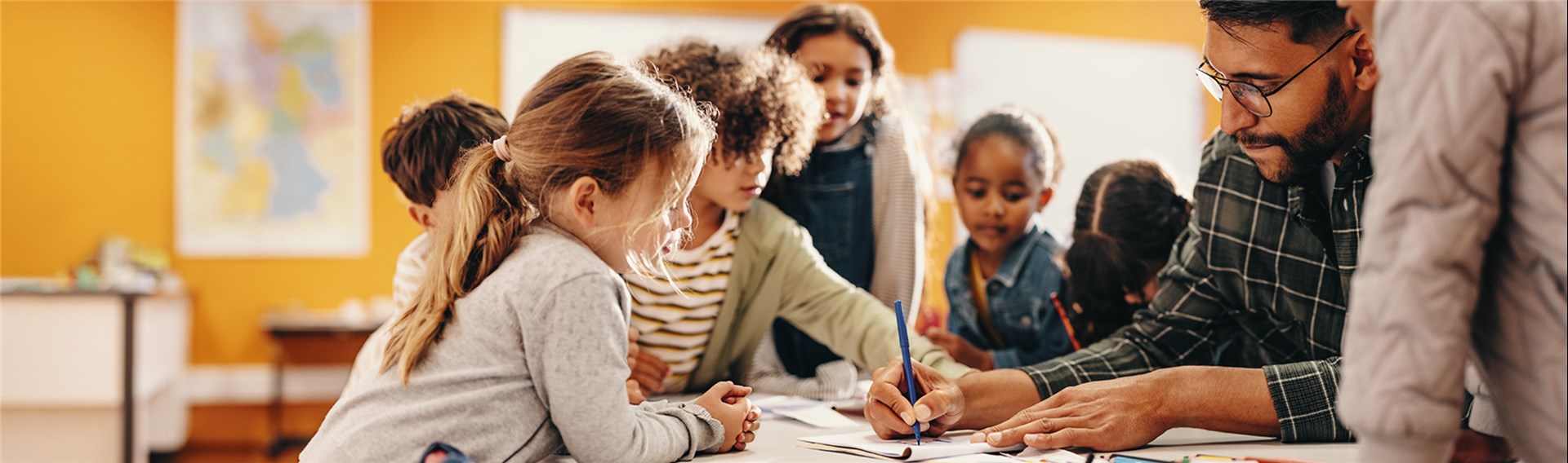 Das Foto zeigt links mehrere Kinder, die einem Erzieher zuschauen, wie er etwas in ein Heft schaut. 