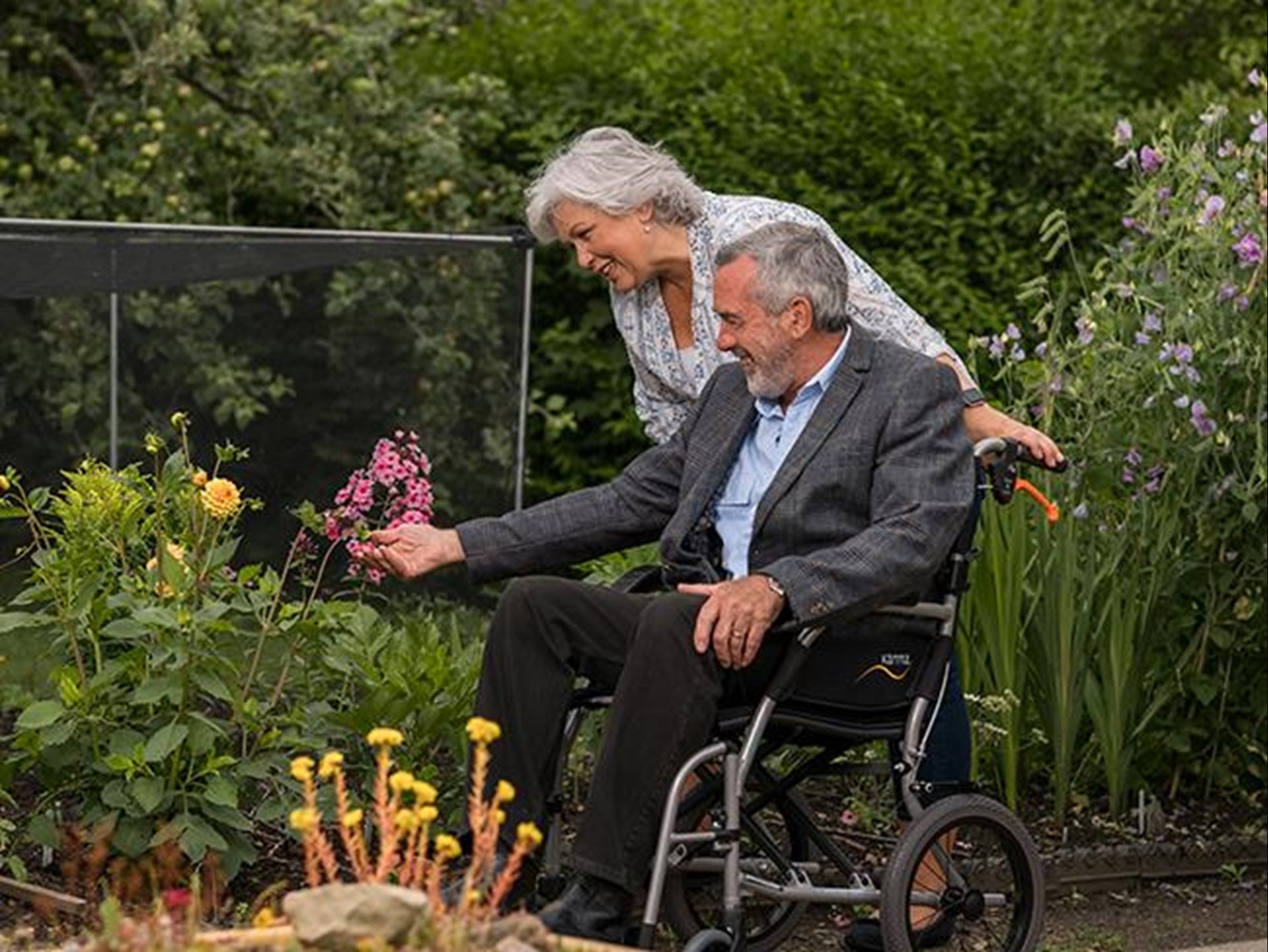 Ein älteres Ehepaar macht einen Spaziergang durch einen Garten. Die Frau schiebt dafür ihren Mann in einem Rollstuhl. Sie halten kurz, um Blumen zu bestaunen. 
