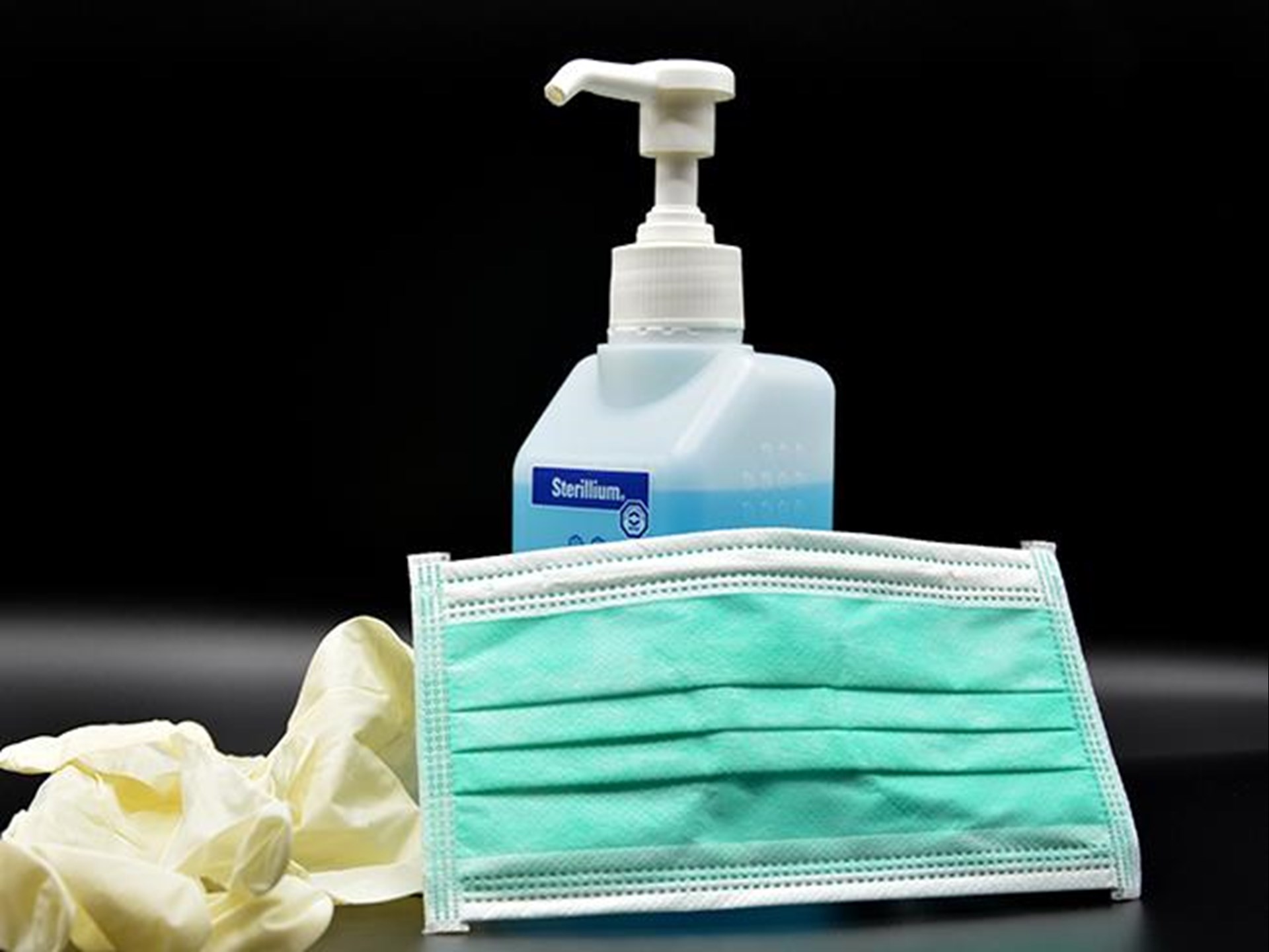 Das Foto zeigt eine Sprühflasche mit Desinfektionsmittel, einen Mund- und Nasenschutz sowie Hygienehandschuhe aus Gummi. 