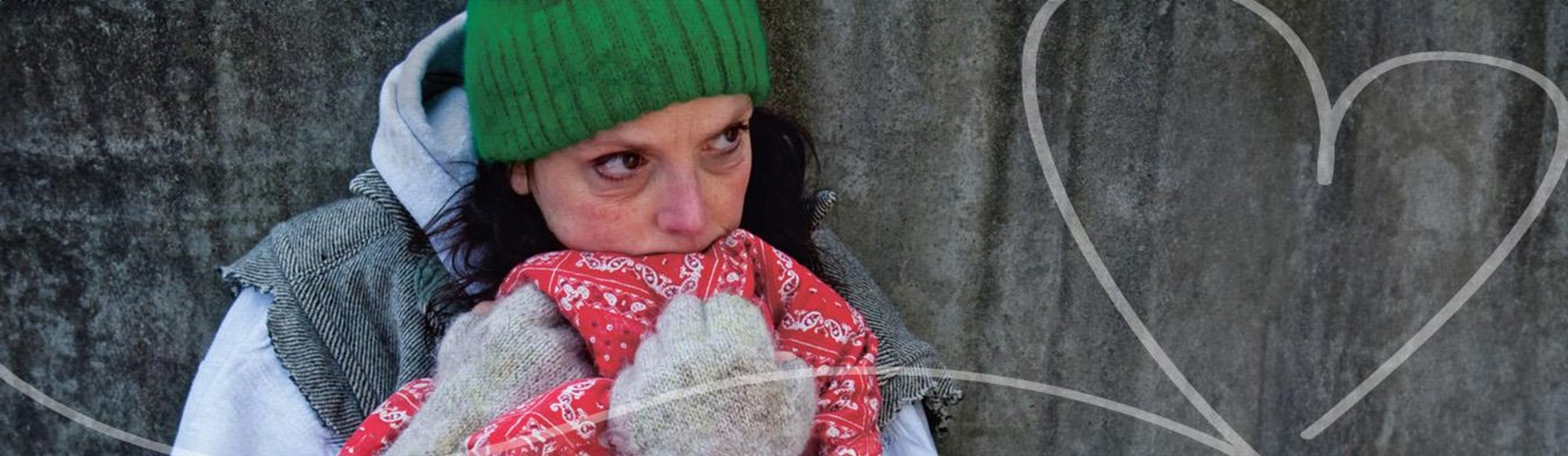Das Foto zeigt eine obdachlose Frau die auf der Straße mit einer roten Decke, grüner Mütze und Handschuhen sitzt