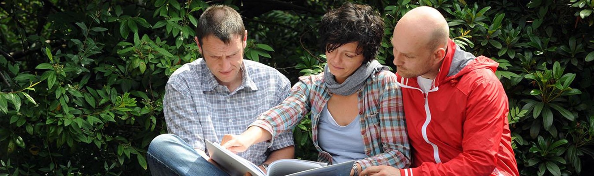 Drei Personen sitzen im Freien und betrachten gemeinsam ein Buch. Eine Frau in einem karierten Hemd und grauem Schal zeigt auf eine Seite,  während die beiden Männer, aufmerksam zuschauen. 